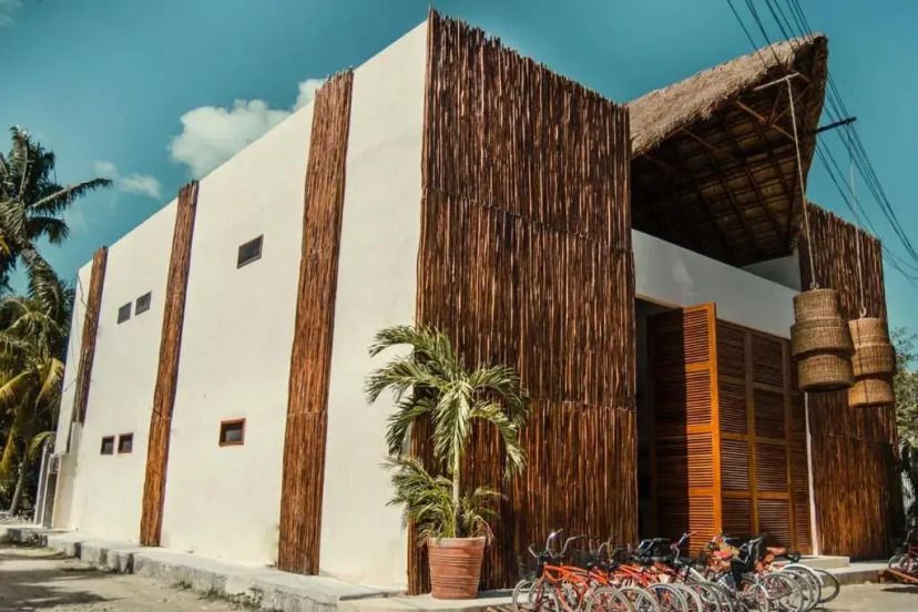 Die Außenansicht des Alcobas Del Mar auf Holbox zeigt ein modernes Gebäude mit rustikalen Holzelementen, großer Eingangshalle und abgestellten Fahrrädern vor dem Eingang unter strahlend blauem Himmel.