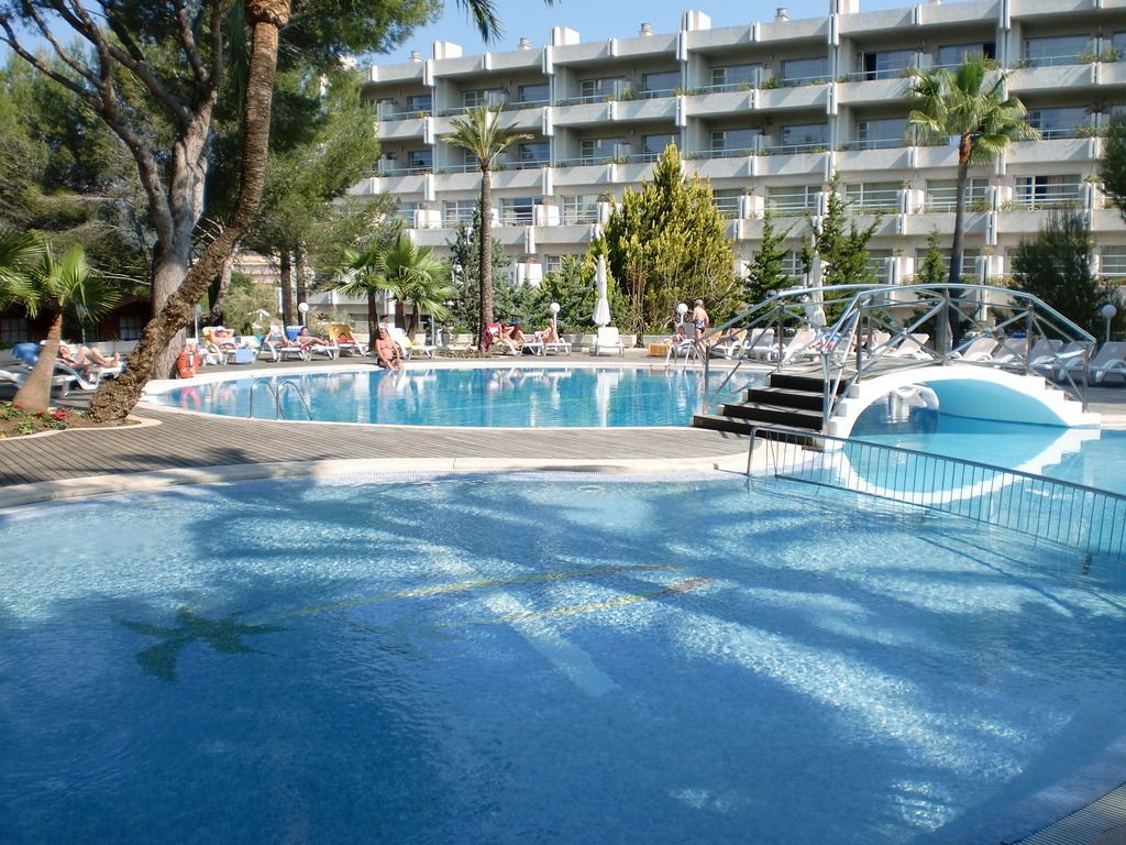Poollandschaft im Allsun Hotel Bella Paguera mit Brücke, Sonnenliegen und umgeben von mediterraner Vegetation – entspannte Atmosphäre unter Palmen.