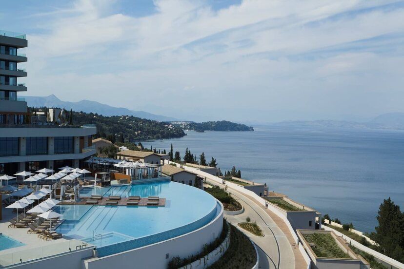 Das Angsana Corfu Resort & Spa mit seinem runden Infinity-Pool und Sonnenliegen liegt auf einer Anhöhe mit weitem Blick über das tiefblaue Meer und die grüne Küste Korfus, umgeben von moderner Architektur und mediterraner Landschaft.