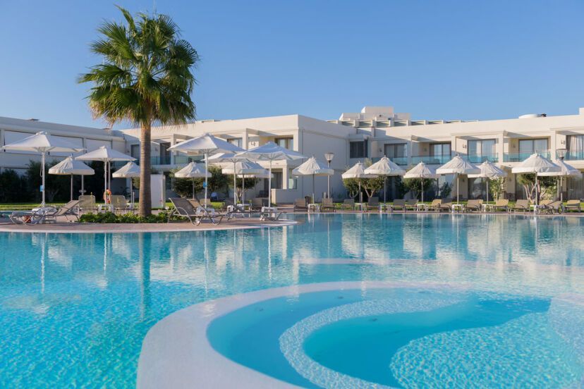 Großer, runder Pool mit Whirlpool-Bereich im Vordergrund des Apollo Blue Hotels, umgeben von Liegen und weißen Sonnenschirmen vor moderner, weißer Hotelanlage.