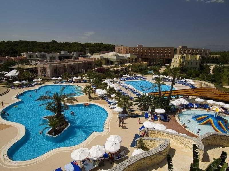 Weitläufige Poollandschaft mit mehreren Becken, Sonnenschirmen und Palmen vor dem Gebäude des Blue Waters Club.