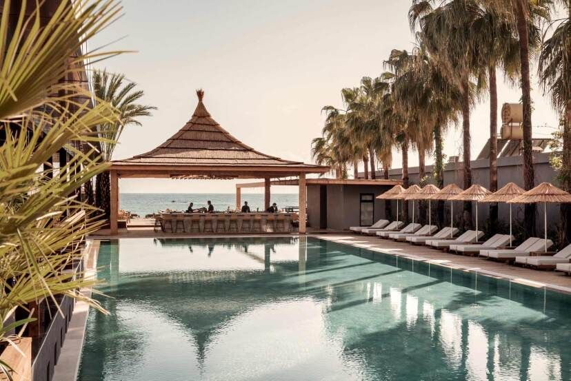 Ein stylischer Poolbereich im Cook’s Club Alanya mit Sonnenliegen, Palmen und einer offenen Bar mit Strohdach – im Hintergrund der Blick auf das Meer bei warmem Abendlicht.