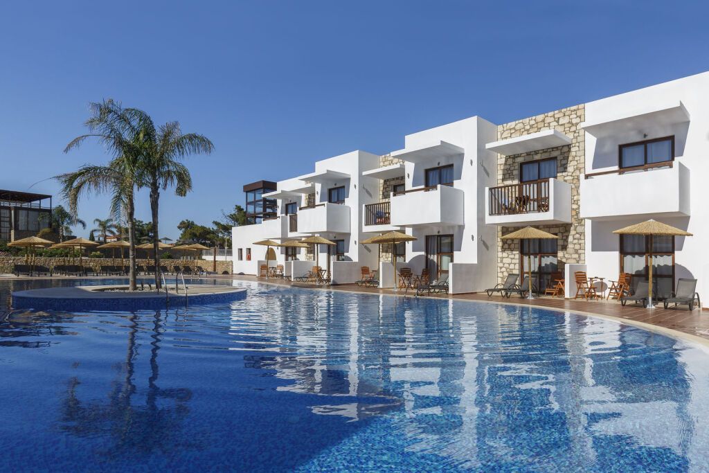Poolbereich des Costa Lindia Blue Star Resort & Spa mit umliegenden weißen Gebäuden im mediterranen Stil, Sonnenliegen, Schirmen und einer kleinen Insel mit Palme.