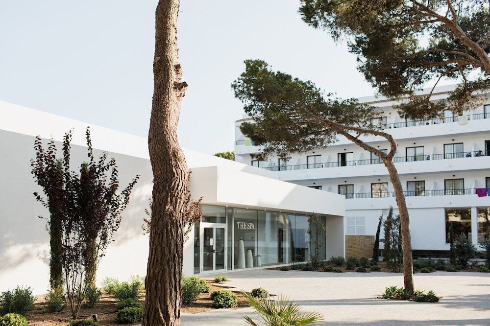 Moderne Hotelanlage mit weißer Fassade und großen Glasfronten, darunter ein Spa-Bereich mit der Aufschrift „The Spa“. Im Vordergrund stehen Pinien und gepflegte mediterrane Pflanzen, im Hintergrund ein mehrstöckiges Gebäude mit Balkonen.