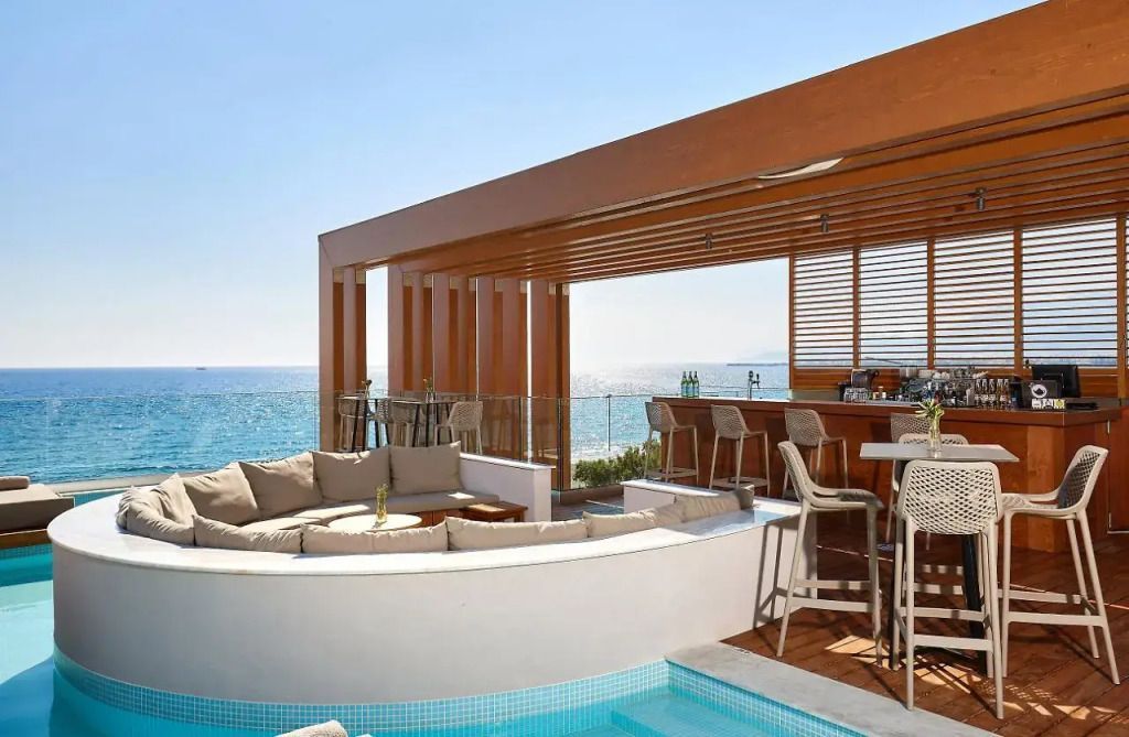 Stylischer Outdoor-Barbereich im Enorme Santanna Beach Resort mit moderner Holzpergola, Rundsofa in einem Pool integriert, Barhockern und Tischen – mit traumhaftem Blick auf das glitzernde Meer.
