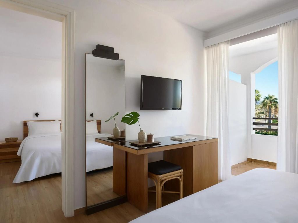 Ein helles, modernes Hotelzimmer im Grecotel Casa Paradiso mit schlichter Einrichtung in Naturtönen. Durch die geöffnete Balkontür ist ein Blick auf Palmen und blauen Himmel sichtbar.