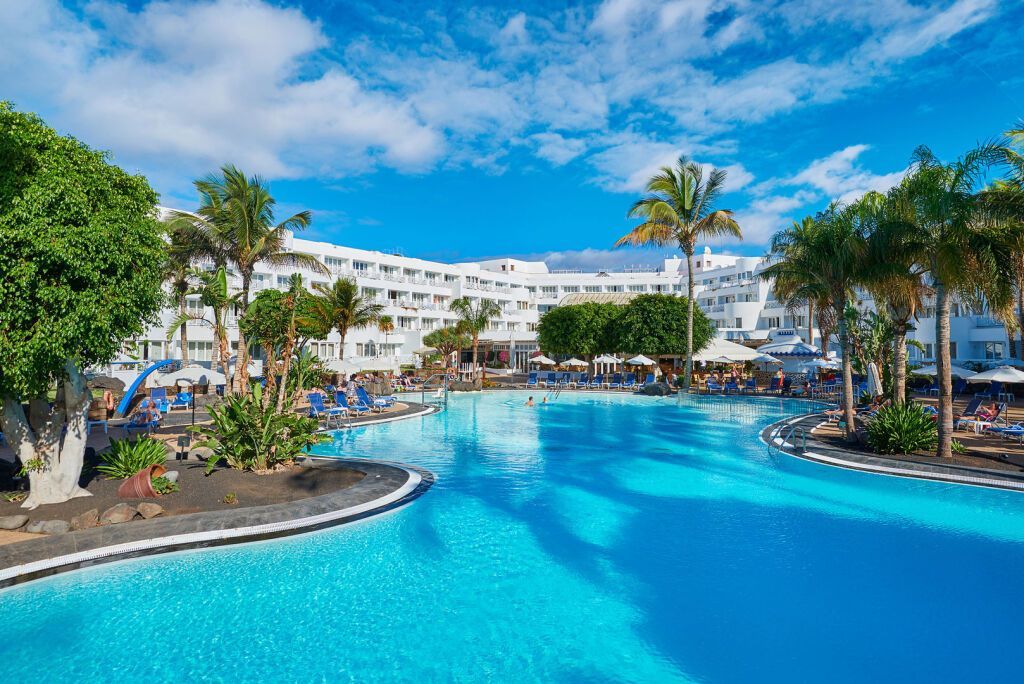 Das Hipotels La Geria auf Lanzarote mit weitläufigem Poolbereich, Palmen, blauen Sonnenliegen und einem weißen Hotelgebäude unter leicht bewölktem Himmel.