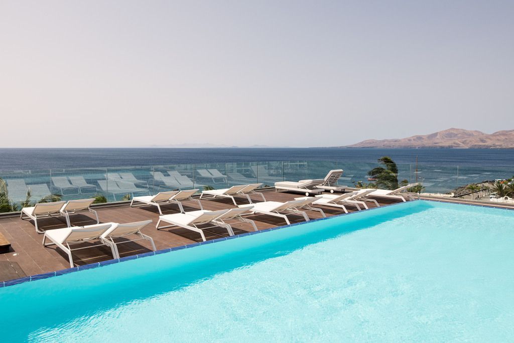 Infinity-Pool des Hotel Fariones mit modernen Liegen, Glasbrüstung und spektakulärem Blick auf den Atlantik und die umliegende Küstenlandschaft bei klarem Himmel.