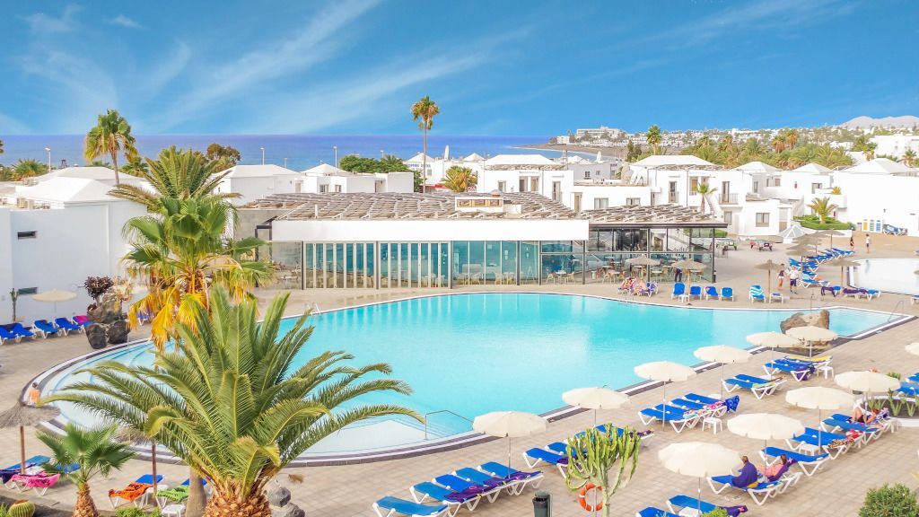 Hotel Floresta auf Lanzarote mit weißer Bungalowanlage, großem Poolbereich, vielen Sonnenliegen und Palmen, im Hintergrund der Atlantik unter blauem Himmel.