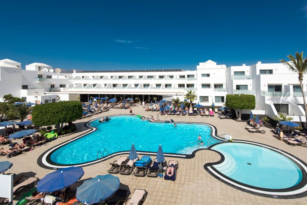 Das Hotel Lanzarote Village mit zentralem Poolbereich, weißen Gebäuden im modernen Stil, Sonnenliegen, Sonnenschirmen und vielen Urlaubsgästen bei strahlendem Himmel.