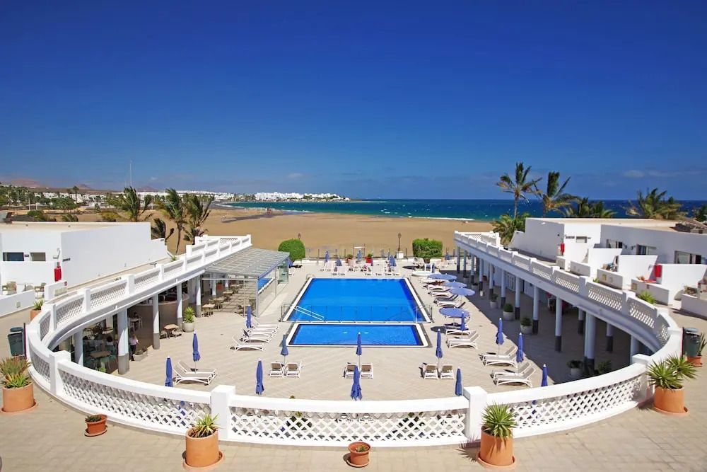 Hotelanlage Las Costas auf Lanzarote mit zentralem Poolbereich, weißen Gebäuden im kanarischen Stil und direktem Zugang zum weitläufigen Sandstrand und Meer.