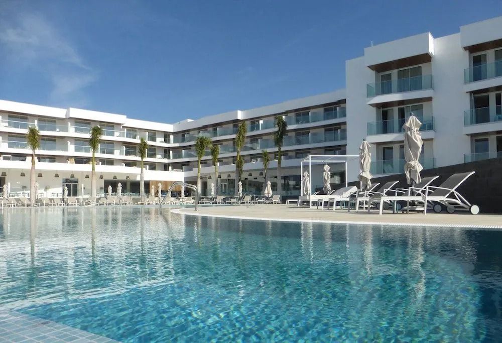 Moderner Poolbereich des Hotel Lava Beach mit weißen Sonnenliegen, Sonnenschirmen und klarer Wasserfläche vor dem weißen Gebäude mit Balkonzimmern.