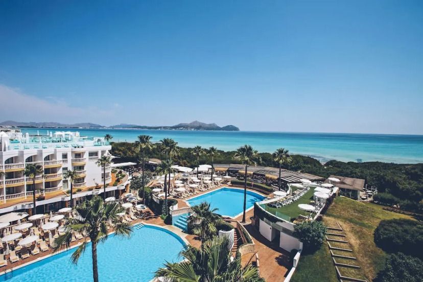 Weißes Hotelgebäude mit mehreren Pools, Sonnenterrassen und Palmen, direkt an der türkisfarbenen Küste von Mallorca mit Blick auf das Meer und die Bucht.