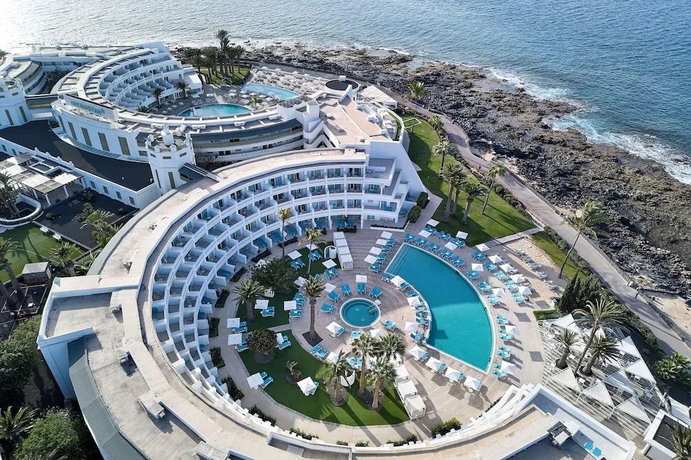 Luftaufnahme des Iberostar Selection Lanzarote Park mit halbkreisförmiger Hotelanlage, Pools im Innenhof, Liegestühlen und direkter Lage an der felsigen Küste mit Meerblick.