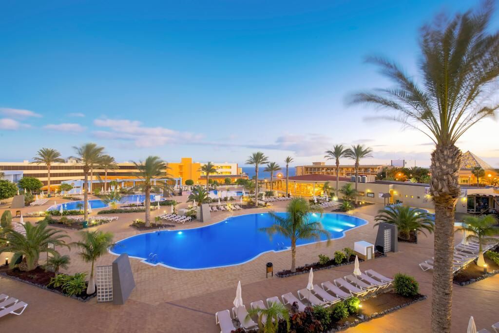 Beleuchteter Poolbereich des Iberostar Waves Gaviotas Park bei Sonnenuntergang mit Palmen, Liegen und Blick aufs Meer im Hintergrund.