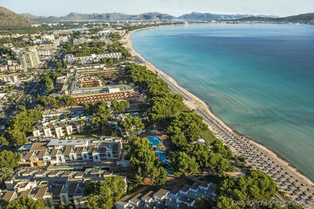 Luftaufnahme des Iberostar Waves Playa de Muro Resorts auf Mallorca mit direkter Strandlage an einer langen, feinsandigen Bucht, zahlreichen Sonnenschirmen am Wasser, weitläufigen Hotelanlagen, Pools und grünen Pinien zwischen den Gebäuden.