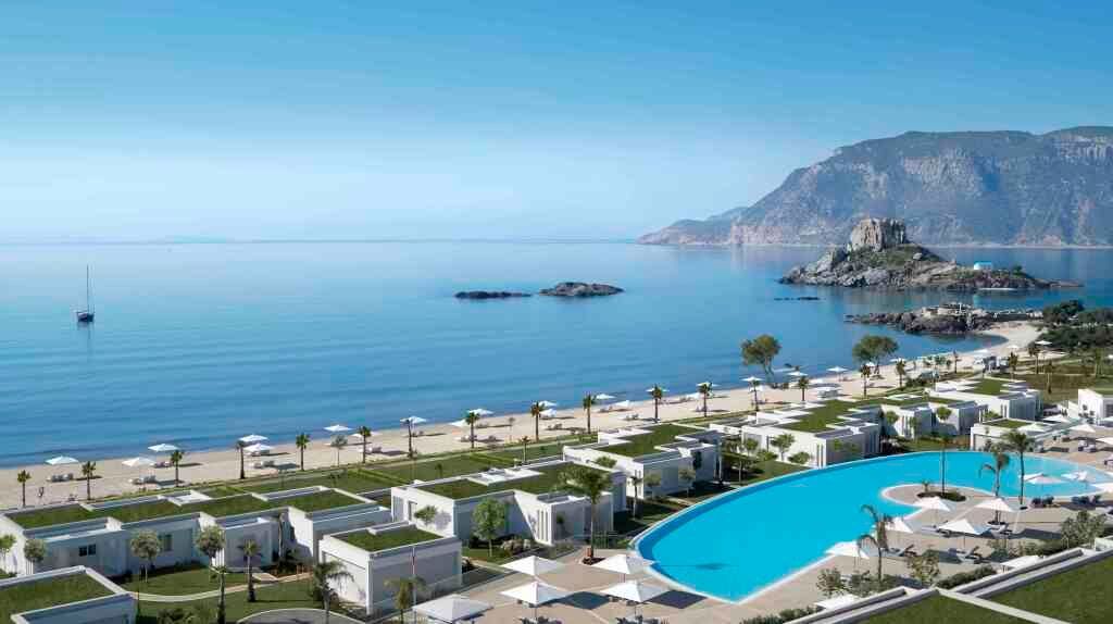 Blick auf das luxuriöse Resort Ikos Aria auf Kos mit weißen Villen, großer Poolanlage, Palmen und direktem Zugang zum Sandstrand mit Blick auf das türkisfarbene Meer und eine Felseninsel.