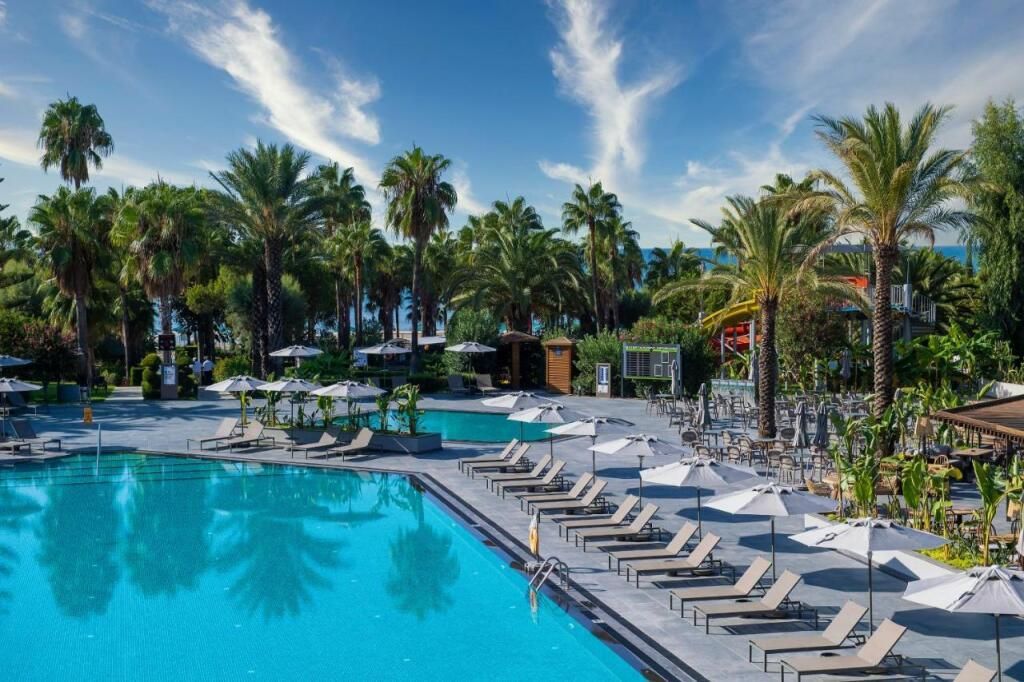 Blick auf die weitläufige Poolanlage des Miramare Beach Hotels mit Palmenallee, Sonnenliegen und direktem Zugang zum Sandstrand am türkisblauen Meer.