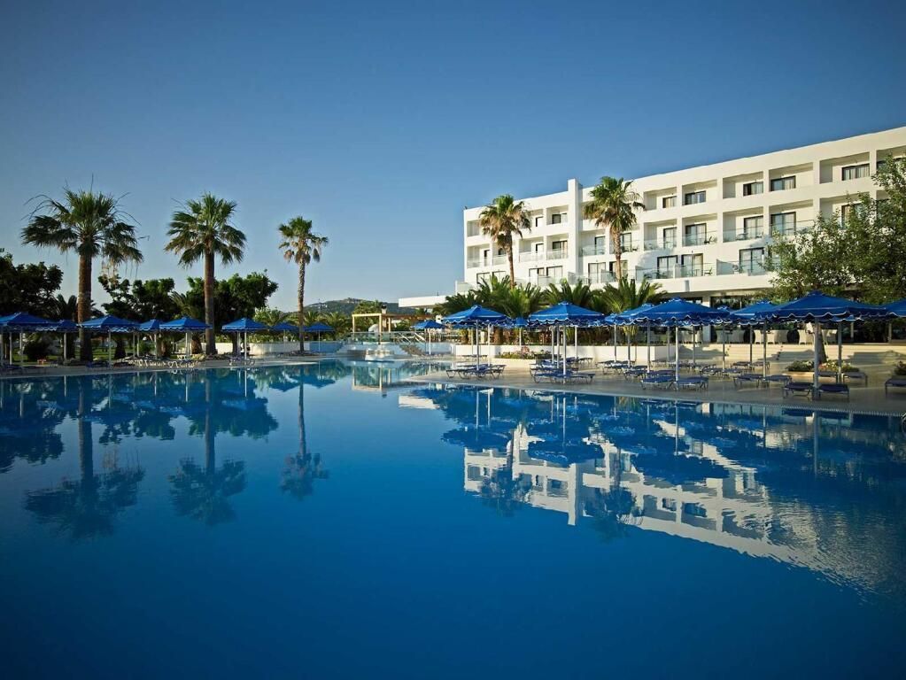 Hotel mit großer Poolanlage, umgeben von blauen Sonnenschirmen, Palmen und einem modernen weißen Gebäude im Hintergrund.