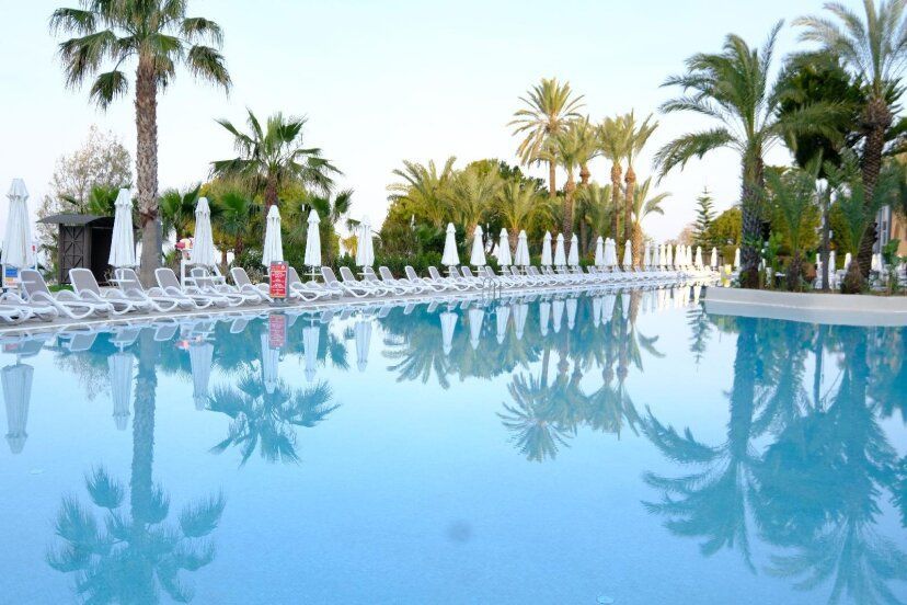 Großer Außenpool eines Resorts an der Türkischen Riviera, umgeben von zahlreichen weißen Sonnenliegen und Sonnenschirmen. Palmen spiegeln sich im ruhigen Wasser des Pools, das eine entspannte Urlaubsatmosphäre vermittelt.