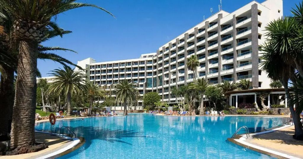 Großer Pool mit Palmen im Vordergrund und dem mehrstöckigen Gebäude des Paradisus by Meliá Fuerteventura im Hintergrund bei blauem Himmel.