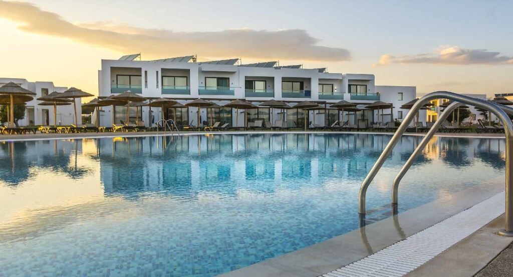 Großer Pool des TUI MAGIC LIFE Plimmiri mit modernen, weißen Gebäuden im Hintergrund und Liegen mit Strohdachschirmen bei Sonnenuntergangsstimmung.