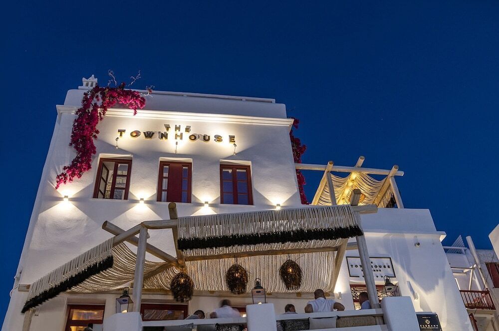 Beleuchtete Fassade des The TownHouse Mykonos bei Nacht mit klassisch weißer Kykladen-Architektur, roten Fensterläden, Dachterrasse und blühender Bougainvillea – stilvoll und einladend.