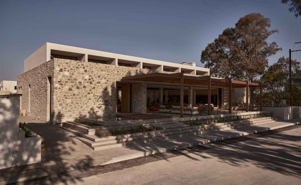 Steinverkleidetes Gebäude mit moderner Architektur, großen Fenstern und überdachter Terrasse mit Sitzgelegenheiten, umgeben von Bäumen und minimalistisch gestalteten Stufen am Eingang.