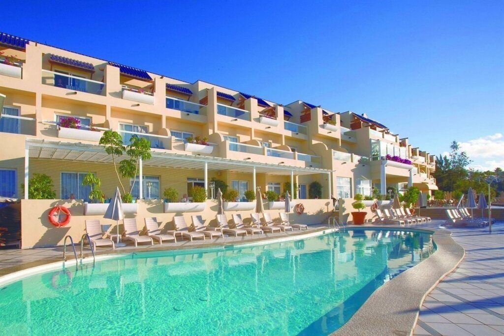 Außenansicht des Hotels XQ El Palacete mit mehreren Etagen und Balkonen, davor ein Pool mit Sonnenliegen und Sonnenschirmen unter strahlend blauem Himmel.