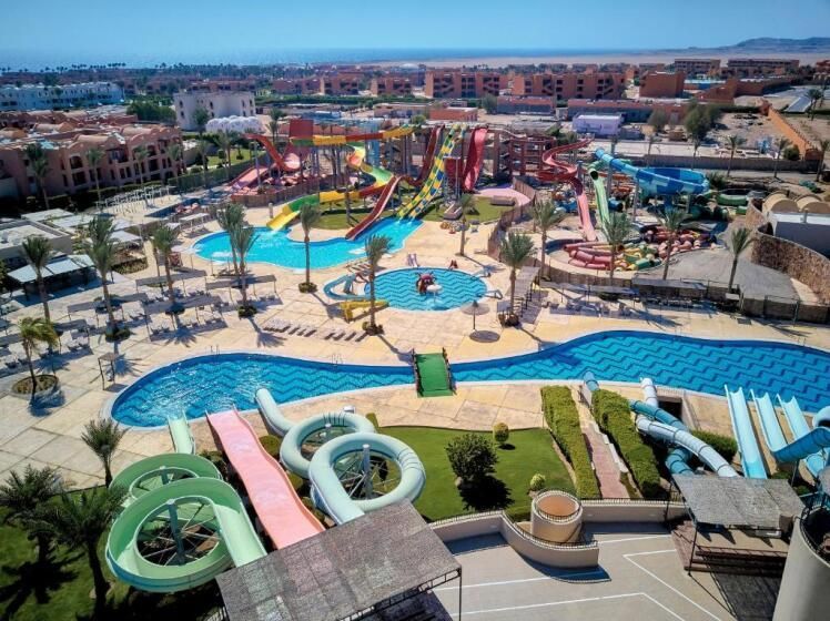 Luftaufnahme des JAZ Elite Maraya Resorts mit großem Wasserpark, bunten Rutschen, mehreren Pools, Palmen und Liegeflächen, umgeben von Gebäuden in Wüstenfarben und Blick bis zur Küste.
