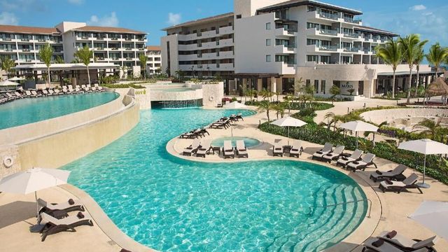 Poolbereich des Dreams Playa Mujeres Golf & Spa Resort mit geschwungenen Becken, Sonnenliegen und Palmen vor dem modernen Hotelgebäude bei strahlend blauem Himmel.