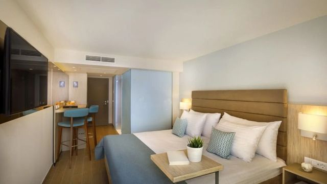 Modern eingerichtetes Hotelzimmer im Girandella Resort mit gemütlichem Doppelbett, hellen Holzmöbeln, stilvollen Deko-Elementen und einem kleinen Sitzbereich mit zwei blauen Barhockern.