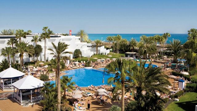 Weitläufige Poollandschaft des Hotel Riu Paraiso Lanzarote mit zahlreichen Sonnenliegen, Palmen und weißen Pavillons, im Hintergrund das weiße Hotelgebäude und der Blick auf das blaue Meer.