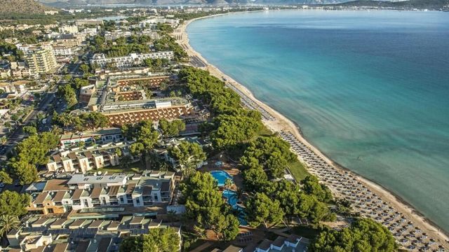 Luftaufnahme des Iberostar Waves Playa de Muro Resorts auf Mallorca mit direkter Strandlage an einer langen, feinsandigen Bucht, zahlreichen Sonnenschirmen am Wasser, weitläufigen Hotelanlagen, Pools und grünen Pinien zwischen den Gebäuden.